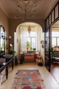 Custom home builder in Westfield exhibits master bath with eclectic mix of styles and a copper tub. There is a red runner in front of a darkspindled wood counter with 2 white sinks and multiple house plants